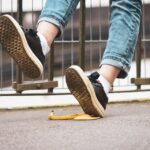 Person in jeans and sneakers about to step on a banana peel on the sidewalk near a fence