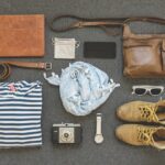 Travel items laid out on gray carpet including striped shirt, brown leather bag, camera, watch, sunglasses, shoes and scarf