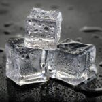 Three clear ice cubes stacked on a wet black surface with water droplets scattered around them