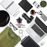 A flat lay of work items on white background including laptop, camera, tablet, notebook, pens, coffee cup, glasses, and green shirt