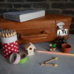 A brown leather suitcase with art supplies scattered around it including colored pencils in a red polka dot cup, washi tape, a tiny birdhouse, and small wooden tools on a gray surface.