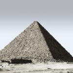A large ancient stone pyramid with rough weathered blocks stands in a desert with sandy ground and clear gray sky