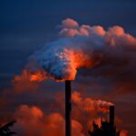 Two tall factory smokestacks releasing thick clouds of steam and smoke into a colorful orange and blue evening sky