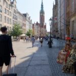 A busy cobblestone street in an old European city with colorful buildings, a tall clock tower, and people in fancy costumes sitting on a bench