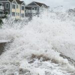 Large ocean waves crash and splash high against a seawall near colorful beach houses during a storm