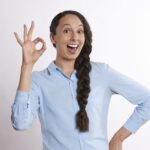 A smiling woman with long braided hair wearing a light blue shirt makes an OK hand sign with her fingers