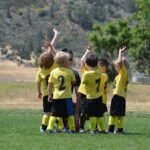 Young soccer players in bright yellow jerseys celebrating together with their arms raised up in victory on a grass field