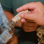 A person's hand putting folded paper money into a clear glass jar, with more money visible inside the jar.