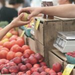 Two hands passing a green apple at a farmers market with red apples, scale, and colorful fruit baskets
