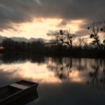 A small wooden boat sits in calm water at sunset, with bare trees reflected in the water and dramatic orange clouds in the sky.