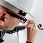 A scientist in a white hard hat looks through a microscope, using his hands to adjust the controls and focus on a sample.