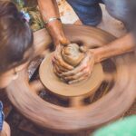 A potter's hands shape wet clay on a spinning pottery wheel while a child watches and learns the craft