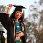 A smiling young woman in black graduation cap and gown holds her diploma up high, wearing a colorful striped stole
