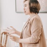 A woman with short brown hair in a tan blazer gestures with her hands while speaking, standing next to a white chair in a bright room.