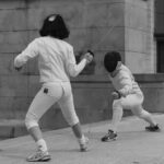 Two people in white outfits and masks practice fencing with thin swords outside a stone building