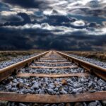 Railroad tracks stretch into the distance under a dramatic cloudy sky, with metal rails and wooden ties on gray gravel