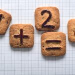 Five square cookies on graph paper with chocolate symbols showing 7, plus sign, 2, equals sign, and 4 to make a math problem