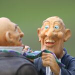 Two people wearing funny masks with exaggerated faces and big smiles appear to be having a playful conversation outdoors