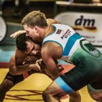 Two wrestlers in colorful uniforms grappling and holding each other during a wrestling match on a yellow and red mat.