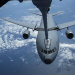 A gray military airplane getting fuel from another plane high above white clouds and blue ocean