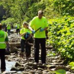 Volunteers in bright yellow-green shirts clean up trash and debris from a rocky creek surrounded by green trees and plants.