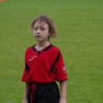 Young child with curly hair wearing a red and black soccer uniform standing on a green grass field