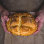 A person's hands gently holding a golden-brown round loaf of bread with a cross pattern on top