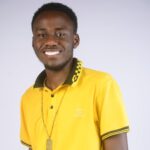 A smiling young Black man wearing a bright yellow polo shirt with green collar and a gold chain necklace against a light background.