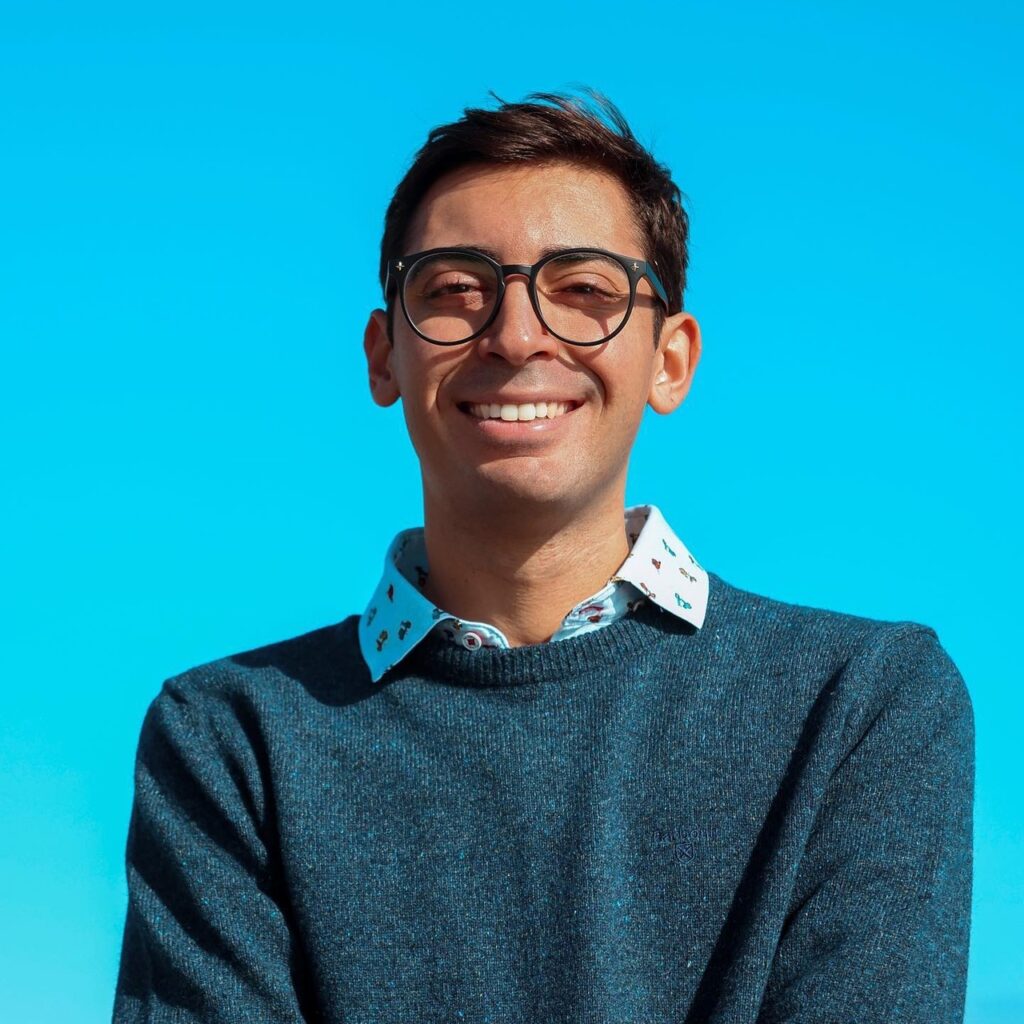 A smiling young man with glasses wearing a blue sweater over a patterned shirt against a bright blue background