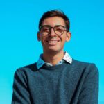A smiling young man with glasses wearing a blue sweater over a patterned shirt against a bright blue background