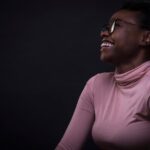 A smiling young woman with glasses and a pink turtleneck looks upward against a dark background, appearing happy and thoughtful.