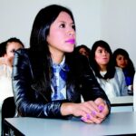 Students sitting at white desks in a classroom, with a girl in a black leather jacket and denim shirt looking up attentively