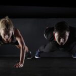 Two people in workout clothes doing push-ups together on a dark gym floor, showing teamwork and exercise.