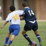 Two soccer players compete for the ball - one in white jersey with number 10, another in dark blue with number 14.