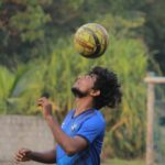 A young soccer player in a blue jersey uses his head to balance a yellow and black soccer ball above him outdoors.