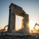Workers using heavy machinery to demolish a large concrete archway with "ABOLISHMENT" written on it during sunset