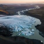 Aerial view of a large white glacier flowing into a muddy river with floating ice chunks, surrounded by brown hills