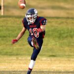 A young football player in a blue Panthers uniform with number 10 runs to catch a football flying through the air on a grass field.