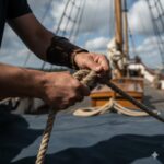 Hands pulling thick rope on sailing ship deck with masts and rigging visible in background on sunny day