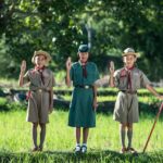 Three young scouts in uniform standing on grass making the scout salute, with one holding a yellow flag and trees behind them.