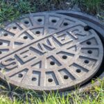 A round metal sewer cover with holes and the word SEWER stamped on it, sitting in grass on the ground