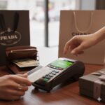 Person using a credit card to pay at a store checkout machine, with shopping bags and wallet nearby on the counter