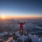 A mountain climber in red gear celebrates with raised ice axes on a snowy peak at sunrise, surrounded by mountains and prayer flags.