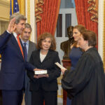 A man in a suit raises his right hand while a woman in black robes holds a book, showing an official swearing-in ceremony.