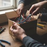 Two people working together to organize colorful wires in a cardboard box, with tools scattered on a wooden table nearby.